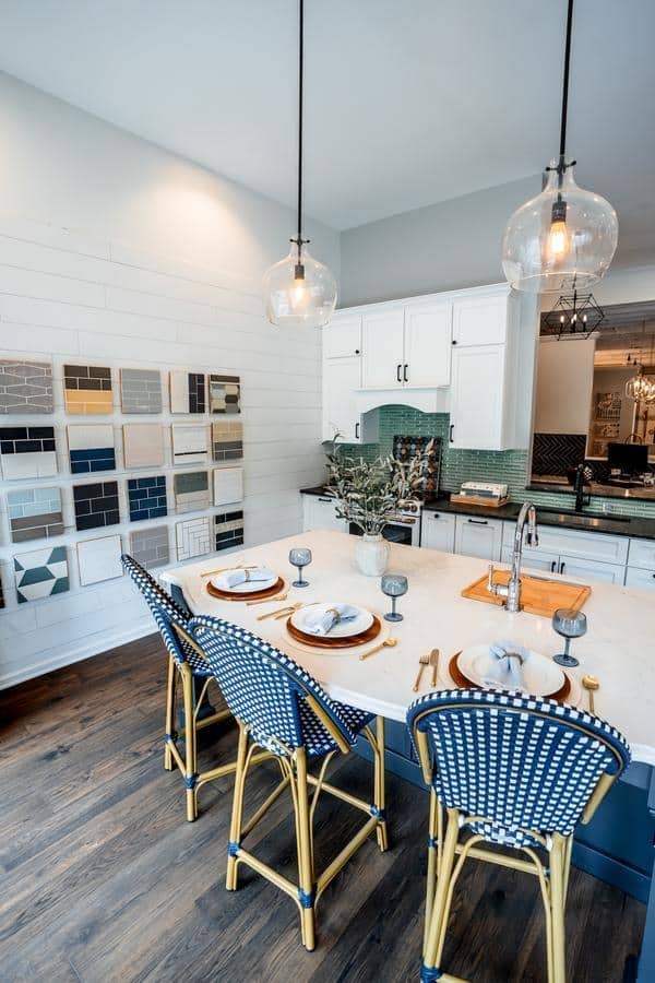 Modern kitchen with a white island set for four, blue and white chairs, pendant lights, white cabinets, green tile backsplash, and a wall decorated with tile samples at the Spencerport Showroom.