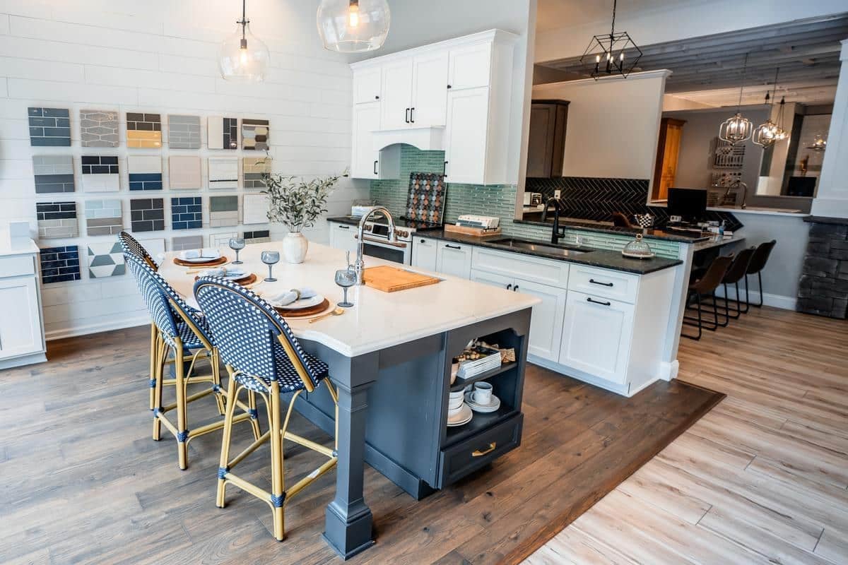 Modern kitchen in the Spencerport Showroom features a large white island, blue and gold barstools, white cabinets, green tile backsplash, wood flooring, and a wall with tile samples. Pendant lights illuminate the island.