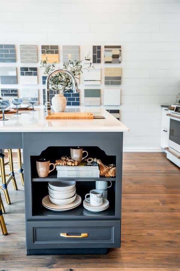 A modern kitchen island with open shelving displaying dishes and mugs, a white countertop with a sink and vase, stools, and sample swatches on the back wall. Visit our Spencerport Showroom to see the wood flooring and oven in the background.