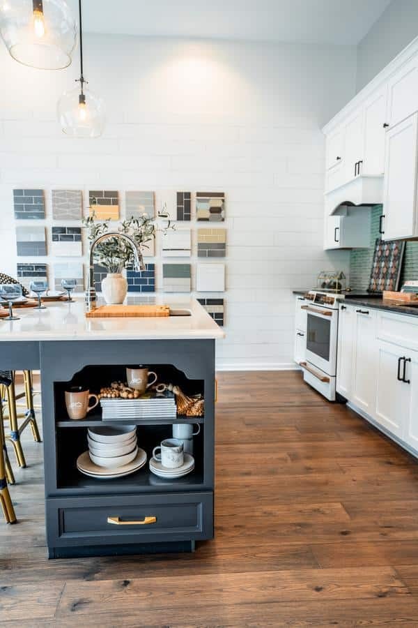 Modern kitchen with white cabinets, wood flooring, and a central island featuring open shelving with cups, plates, and bowls—like those displayed in the Spencerport showroom. Decorative pendant lights hang above a wall of tile samples.