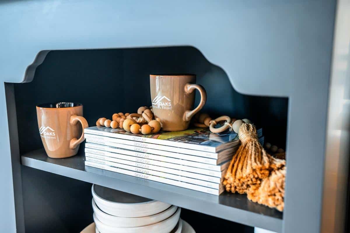 Two brown mugs, a string of wooden beads, a stack of magazines, and decorative tassels are neatly arranged on a dark shelf in the Spencerport Showroom. White plates are stored on the shelf below.