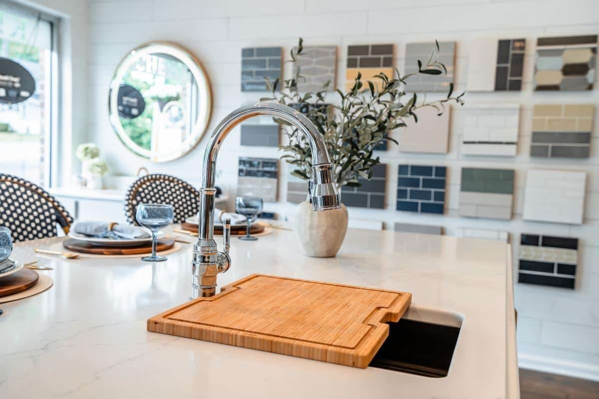 A modern Spencerport Showroom kitchen with a marble island, a shiny chrome faucet, a wooden cutting board over the sink, a vase with branches, and sample tiles displayed on the wall in the background.