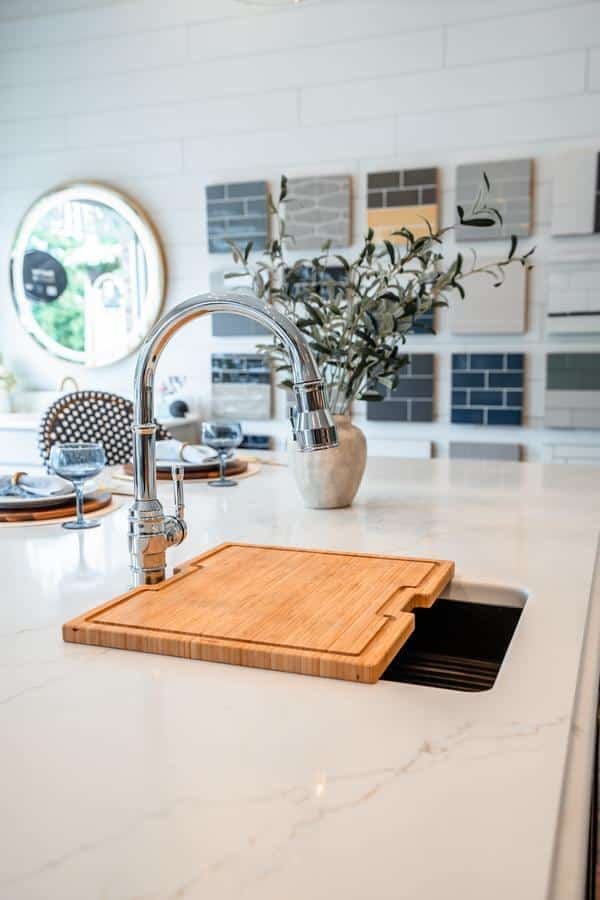 A modern kitchen island with a marble countertop features a chrome faucet, a wooden cutting board over a small sink, a vase with greenery, and sample tiles displayed in the background at the Spencerport Showroom.