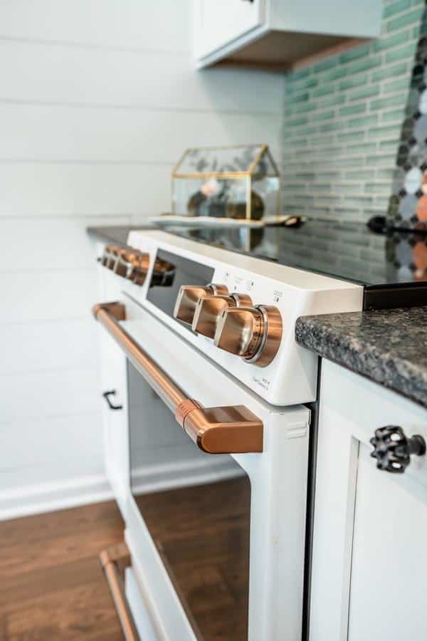 A modern white oven with metallic gold handles and knobs sits in a kitchen with a dark countertop and light green tile backsplash, echoing the style of the Spencerport Showroom. A small decorative glass box is on the counter.