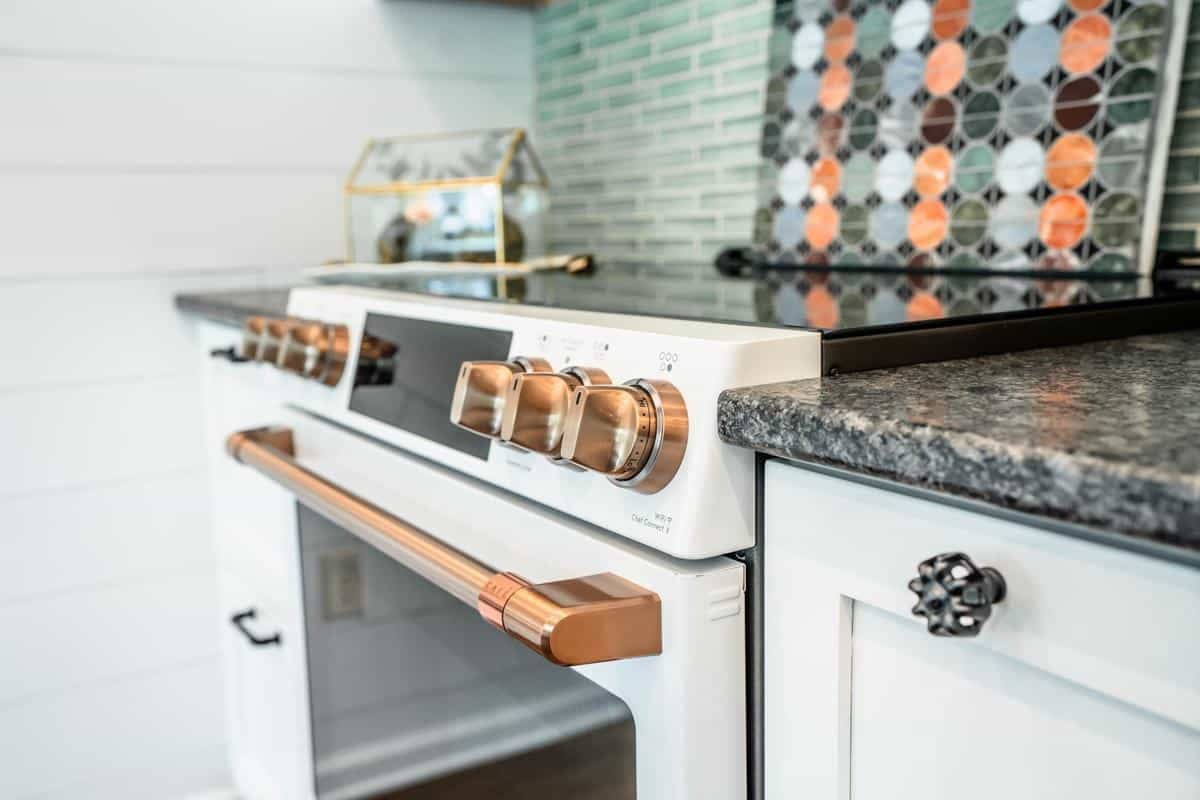 A modern kitchen featuring a white oven with rose gold knobs and handle, a black granite countertop, and a green-tiled backsplash with a colorful circular pattern accent is showcased at the Spencerport Showroom.