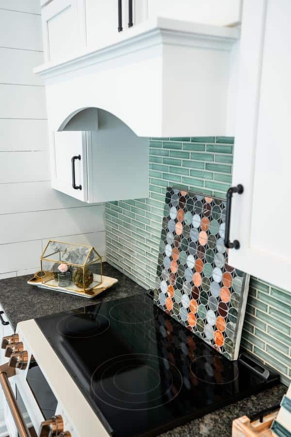 A modern kitchen features a black glass cooktop, white cabinets with black handles, a green glass tile backsplash, and a decorative tray with a geometric terrarium and patterned cutting board—all showcased in the Spencerport Showroom.