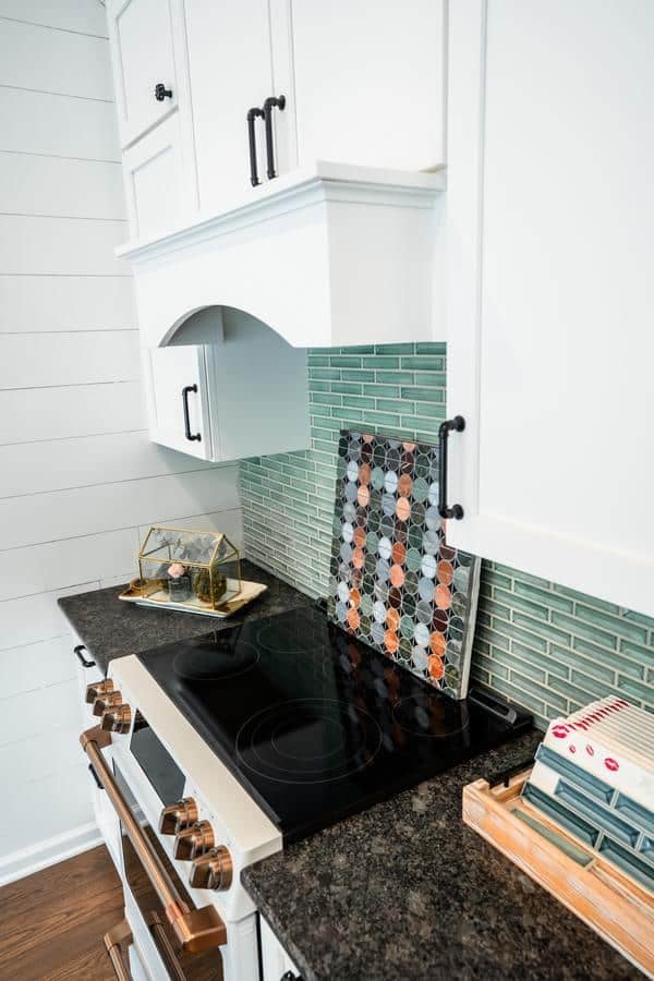 Modern kitchen featuring white cabinets, a black electric stovetop, green glass tile backsplash, and a dark marble countertop with decor items—all inspired by displays at the Spencerport Showroom.