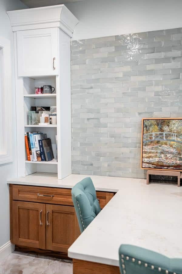 A home office corner from the Spencerport Showroom features a white marble desk, wooden cabinets, a teal upholstered chair, built-in shelves with books and mugs, and a framed nature painting against a light gray subway tile wall.