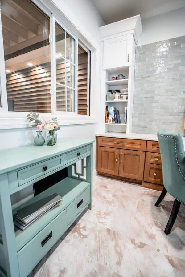 A home office corner inspired by the Spencerport Showroom features a turquoise table, small vase of flowers, wooden cabinets and shelves, a green upholstered chair, and a window overlooking an outdoor area with horizontal siding.