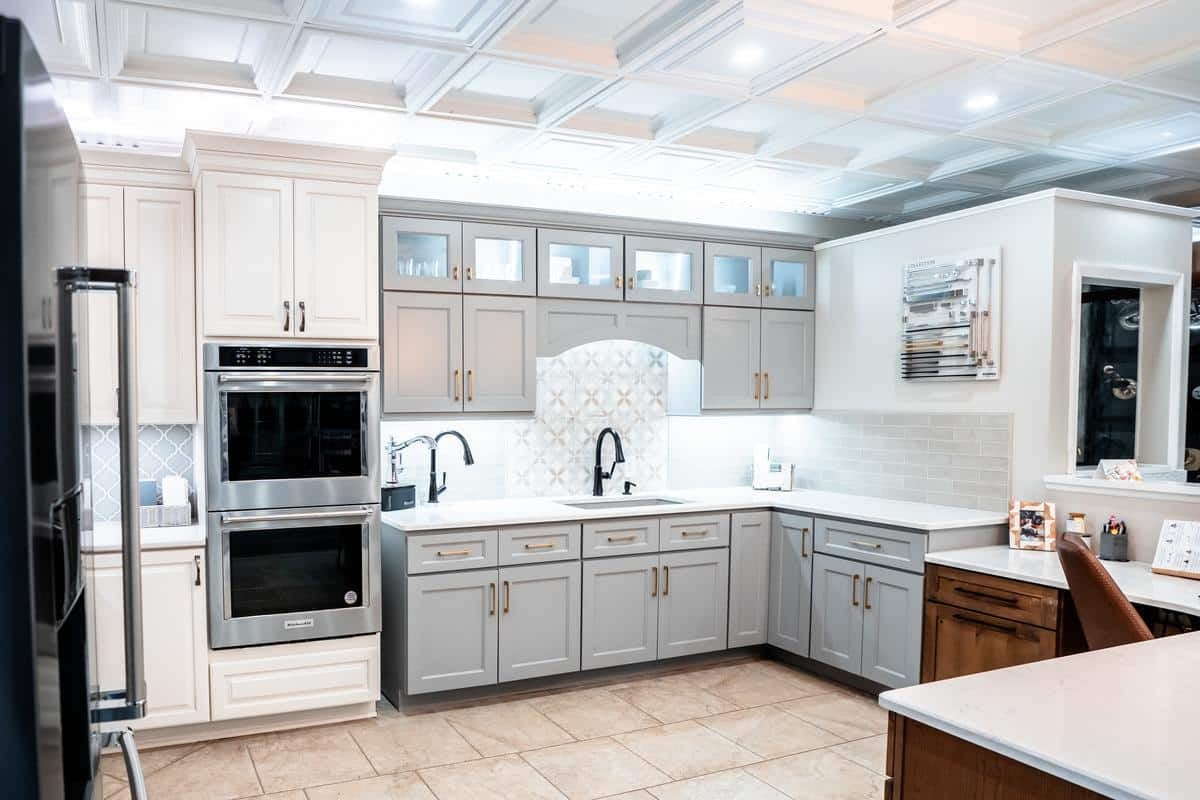 Modern kitchen with white upper cabinets and light gray lower cabinets, built-in double oven, patterned tile backsplash, under-cabinet lighting, and tan tiled floor. Visit our Spencerport Showroom for clean white countertops and a sleek look.
