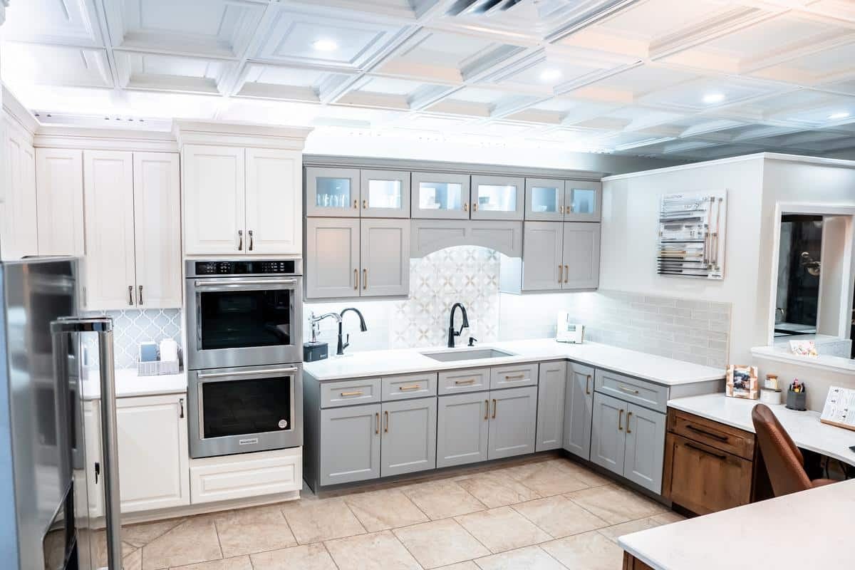 Modern kitchen with white upper cabinets, gray lower cabinets, stainless steel appliances, a patterned backsplash, under-cabinet lighting, and a tile floor. This bright, neatly organized space is featured in our Spencerport Showroom.