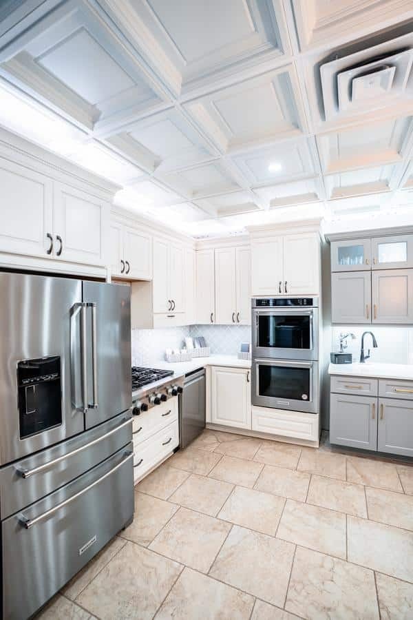 Modern kitchen with stainless steel fridge, double wall ovens, white and gray cabinets, a gas stove, and tiled floor; coffered ceiling with recessed lighting above—see this stunning design at the Spencerport Showroom.
