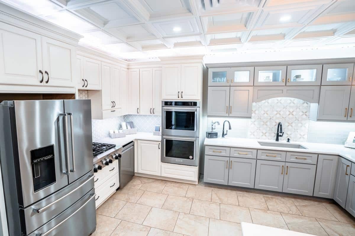 Modern kitchen with white and gray cabinets, stainless steel refrigerator, oven, gas stove, tile backsplash, and a patterned tile accent above the sink—see similar designs at our Spencerport Showroom with decorative panels and recessed lighting.