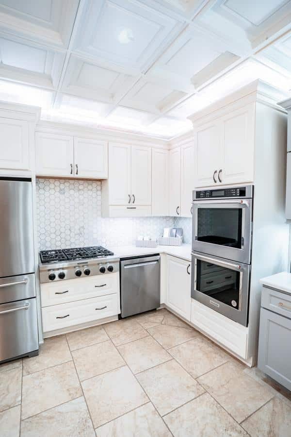 Modern kitchen with white cabinets, stainless steel refrigerator, dishwasher, double wall ovens, gas stove, and a hexagonal tile backsplash. Visit our Spencerport Showroom to see the light tile floors and bright overhead lighting in person.
