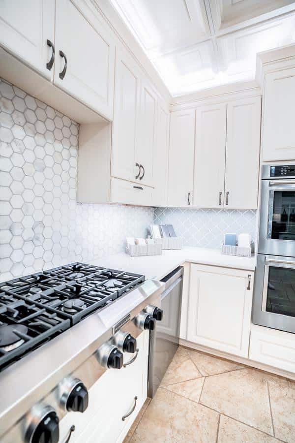 Modern kitchen with white cabinets, stainless steel gas stove and oven, light tile backsplash, beige tiled floor, and under-cabinet lighting—creating a bright, clean space inspired by the Spencerport Showroom.
