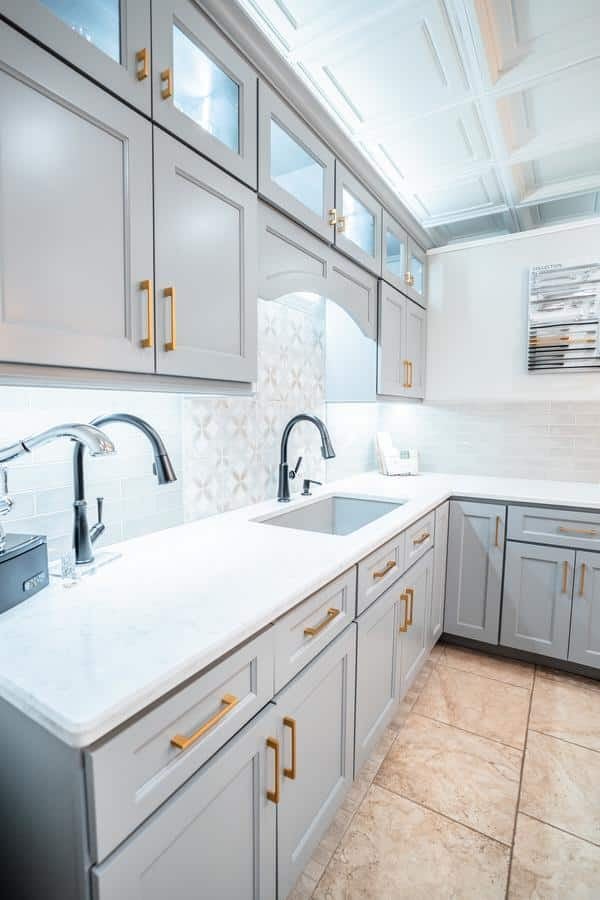 Modern kitchen with light gray cabinets, gold handles, white marble countertops, two sinks with chrome faucets, neutral-toned backsplash, and beige tiled floor—available to view at the Spencerport Showroom. Upper cabinets feature glass panels and under-cabinet lighting.