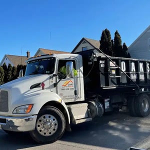 A large white dump truck parked on a suburban street. The truck has a black dumpster attached.