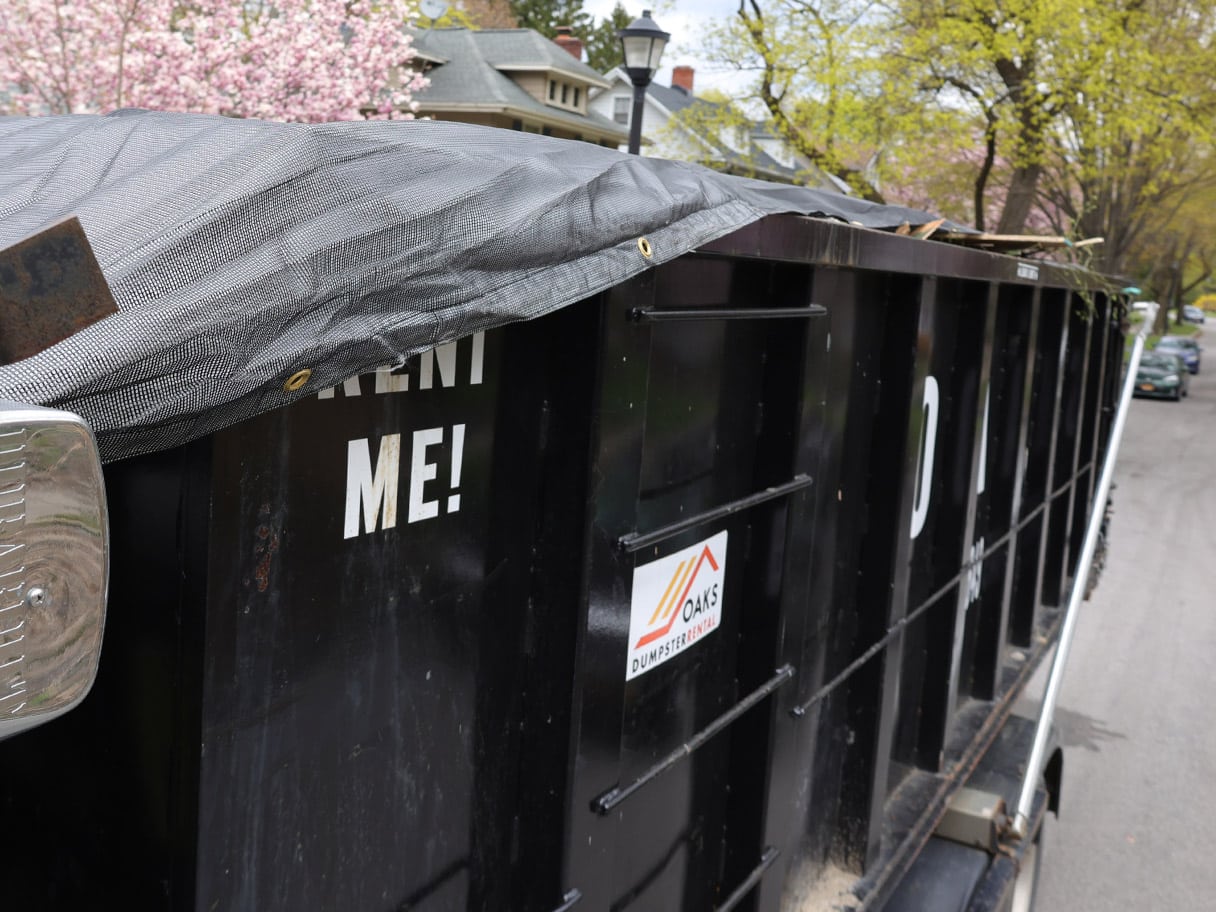 A black dumpster with a gray tarp on top sits on a residential street.