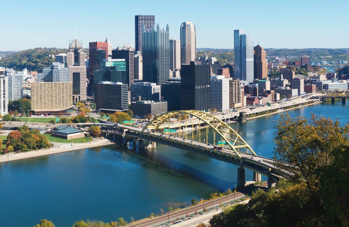 Pittsburgh city skyline with a yellow bridge over a river. The bridge connects the city to a park.