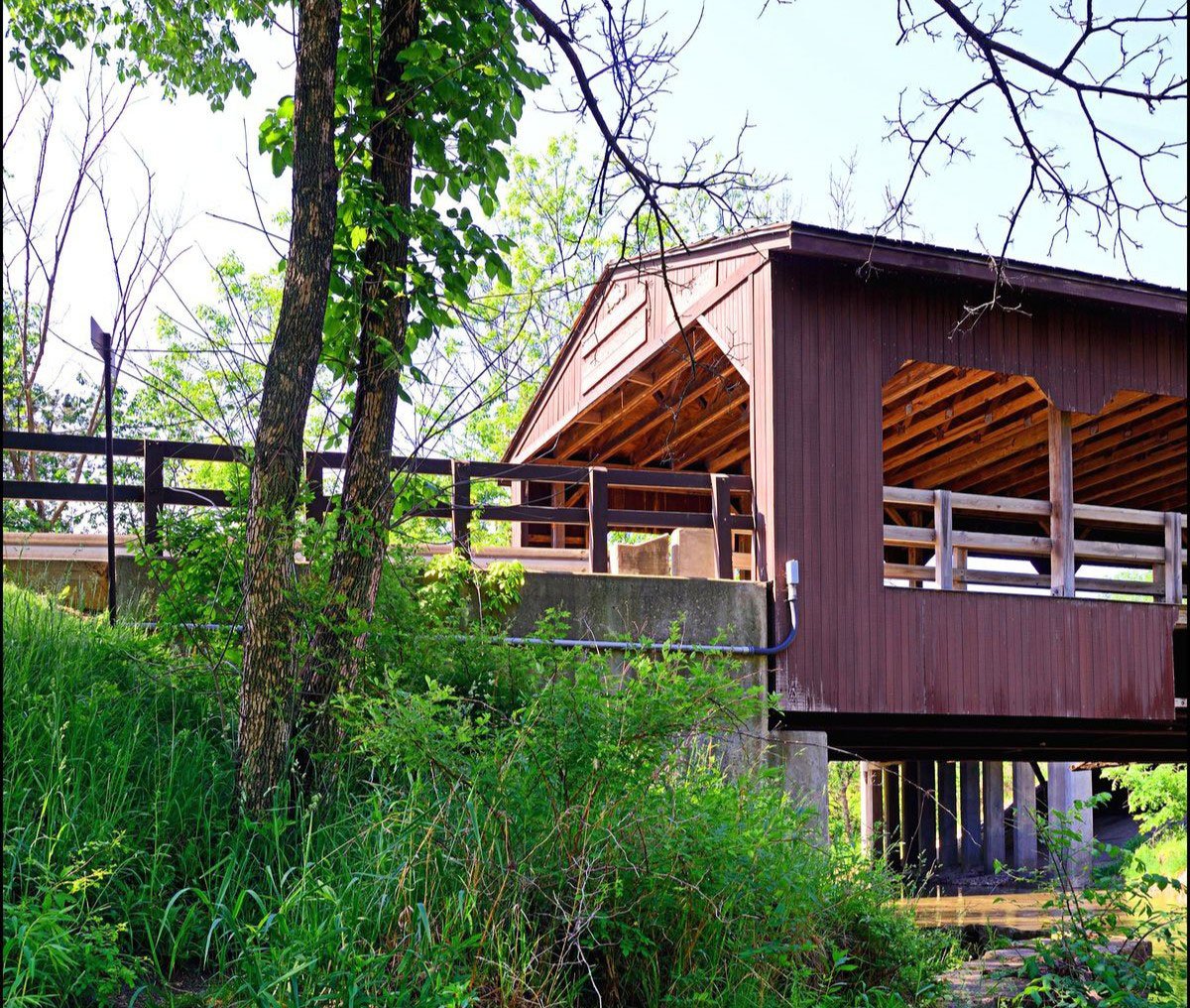 A brown wooden covered bridge with a gable roof surrounded by greenery. The bridge has a railing and appears to be elevated.