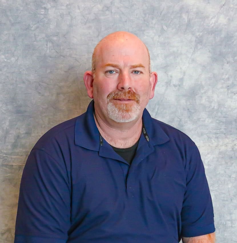 A bald man with a moustache wearing a blue shirt. He is standing against a grey background.