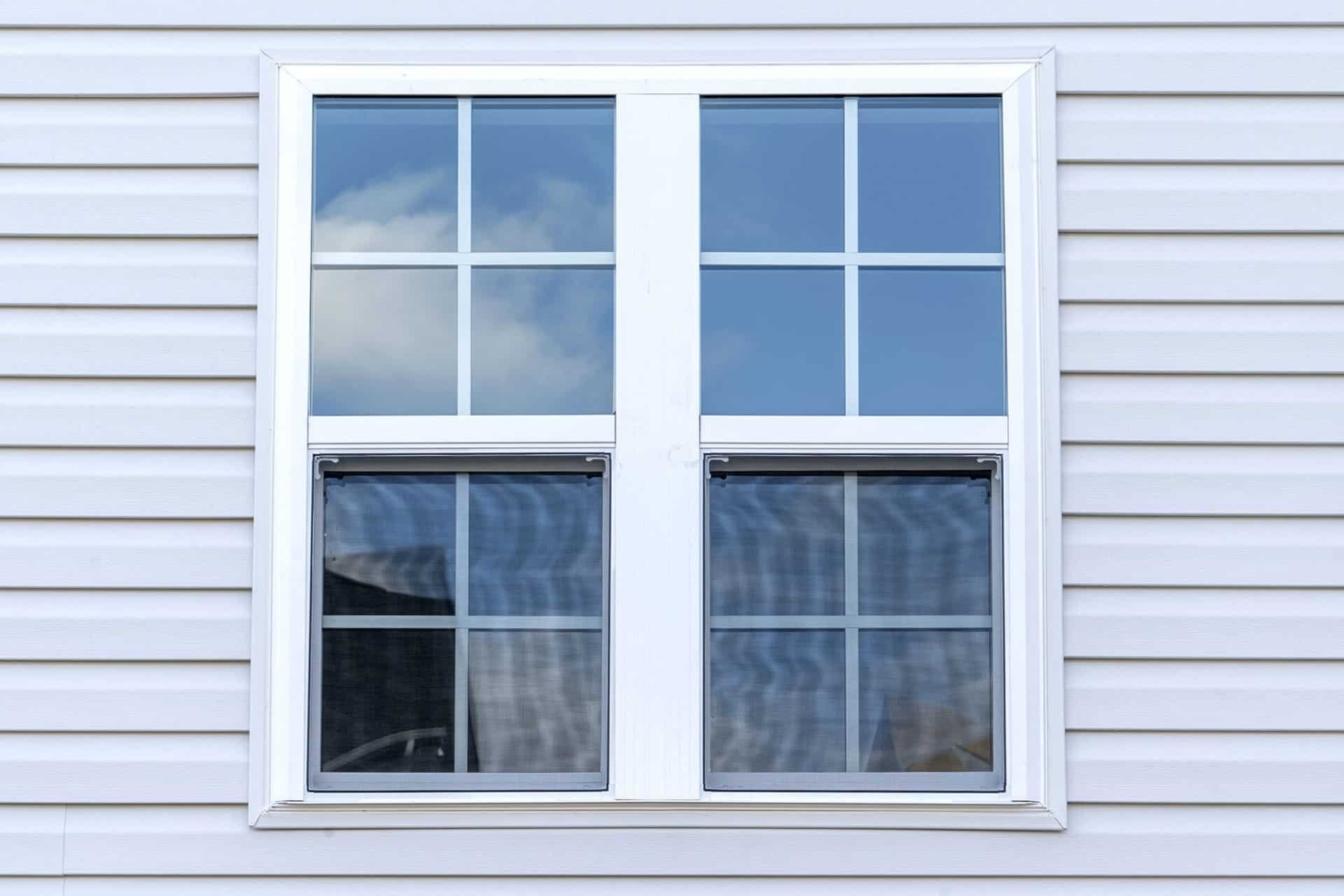 A white double hung window on a gray siding house. The window reflects a blue sky.