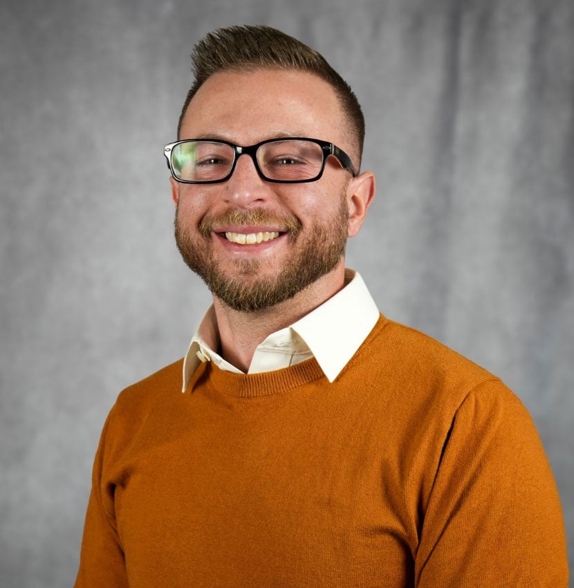 A man with a beard smiles wearing glasses and an orange sweater.