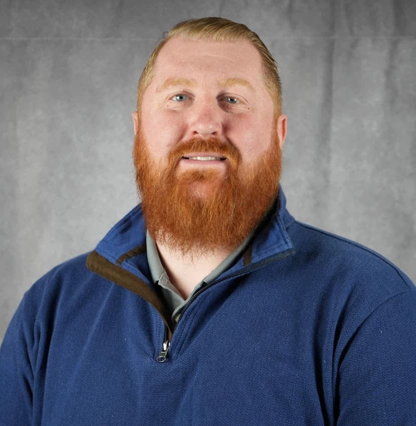 A man with red hair and a beard smiles at the camera. He is wearing a blue sweater.