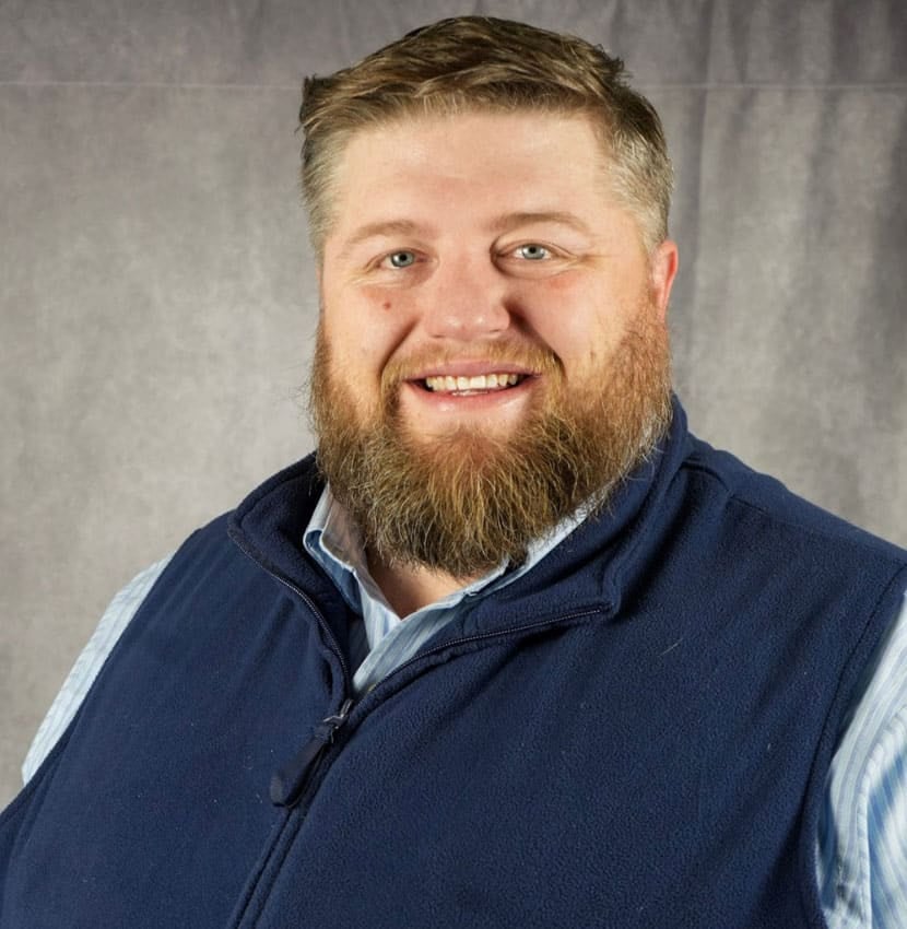 A man with a beard smiles at the camera. He is wearing a blue vest.