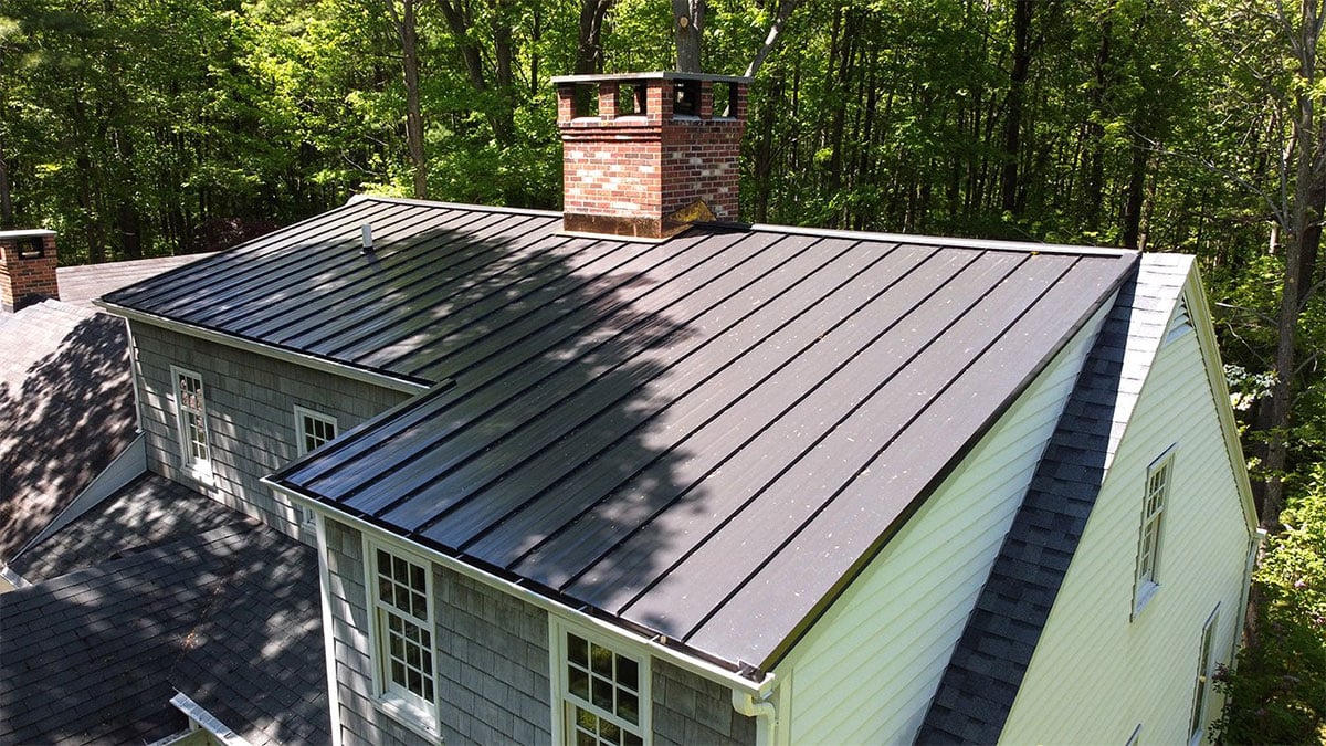 A house with a metal roof and a brick chimney surrounded by trees. The house has siding and multiple windows.