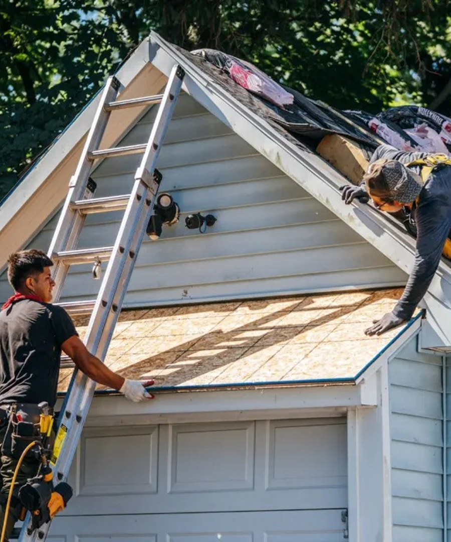 Two workers on a roof replacing shingles. A construction worker on a ladder assists another on the roof.