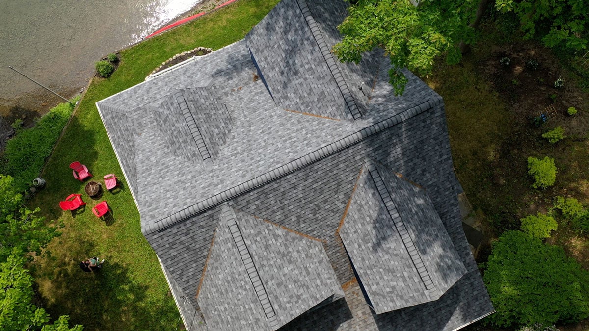 Aerial view of a house with a gray roof surrounded by trees and a lawn with red umbrellas. The house has multiple gable roofs.