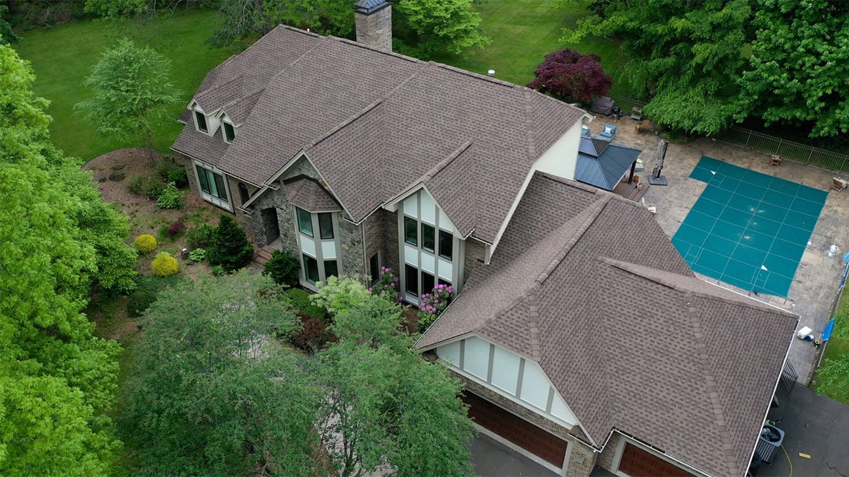 A large house with a swimming pool and lush green garden. The house features a brown roof and multiple windows.