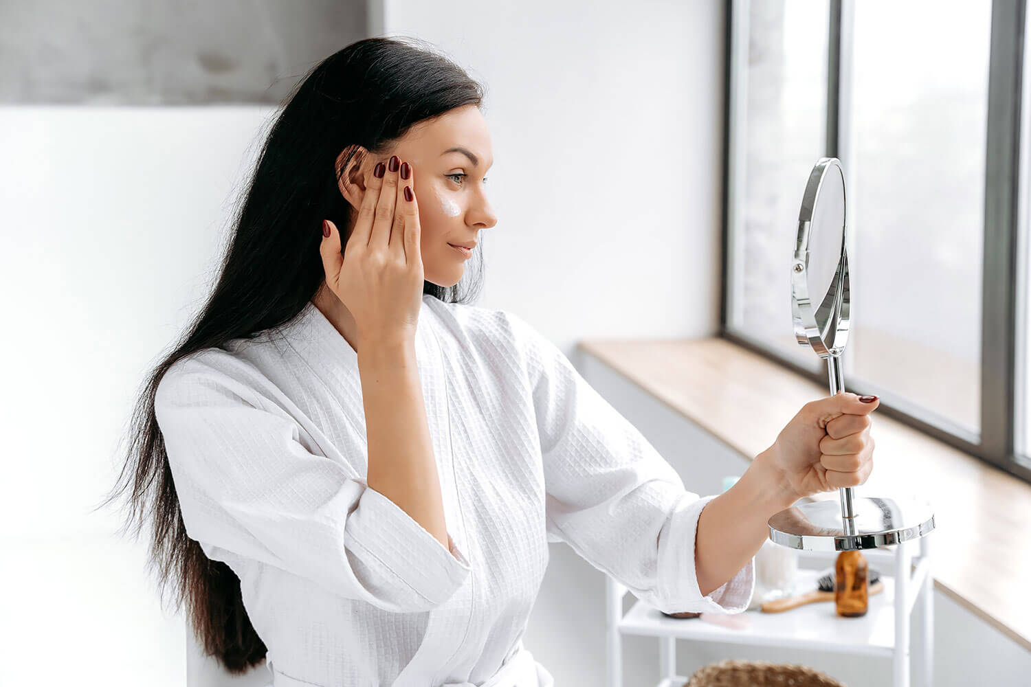 Beauty Blog Creating the Perfect Care Routine for Combination Skin. Happy caucasian woman, in bathrobe, holds a mirror in her hand, applies a moisturizing, anti-aging cream on her face, takes care of her skin and youth, prevents wrinkles, smile.
