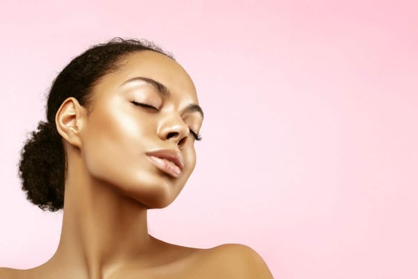 Our Guide on How to Use Makeup Highlighter. African American skincare models portrait. Beauty spa treatment concept. Young girl posing with closed eyes against pink background
