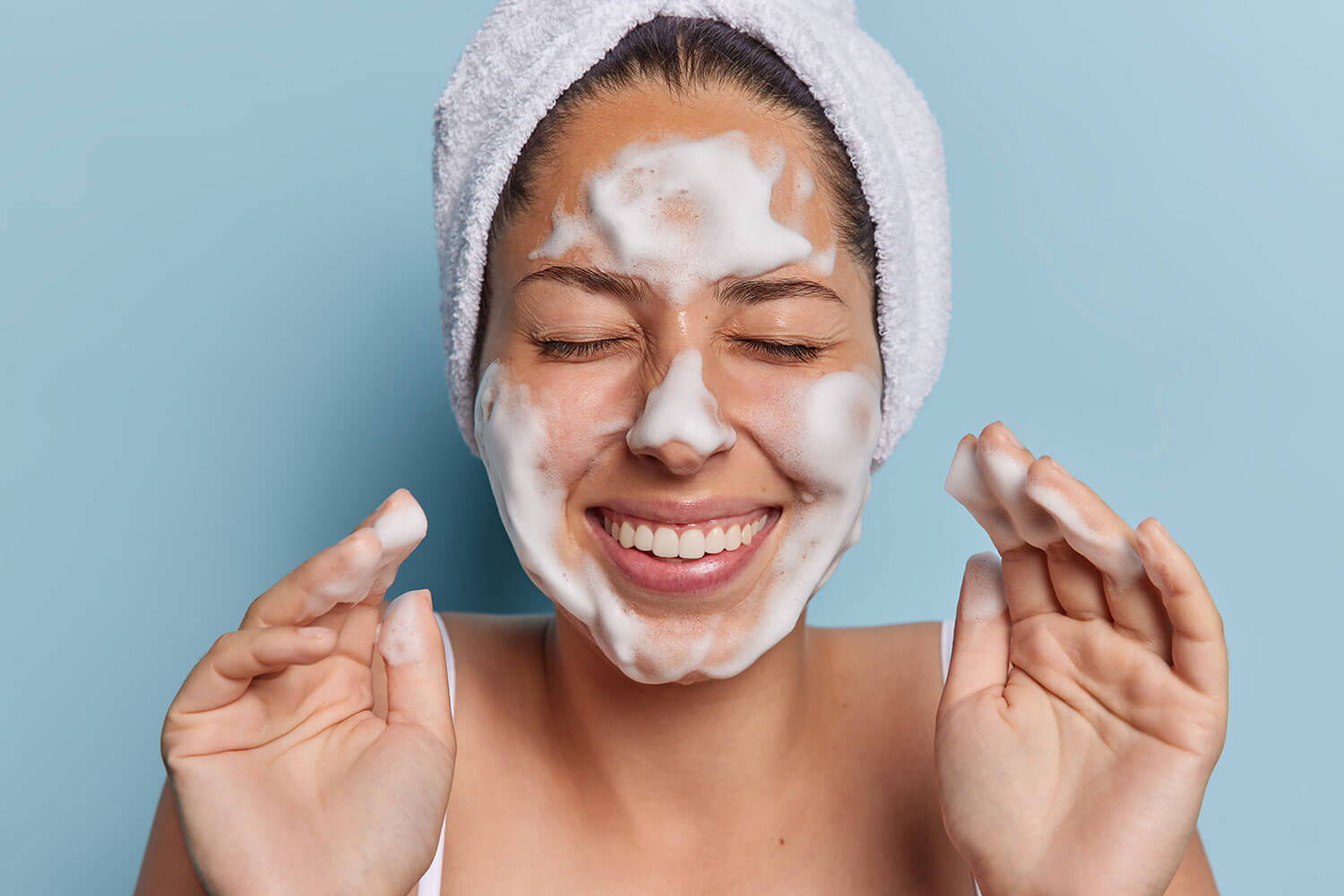 6 Face Washes for Oily Skin to Balance Your Radiance. Headshot of young female model indulges in her skincare routine delicately applies foam to her cheeks revealing radiant complexion wears bath towel on head giggles positively isolated over blue wall