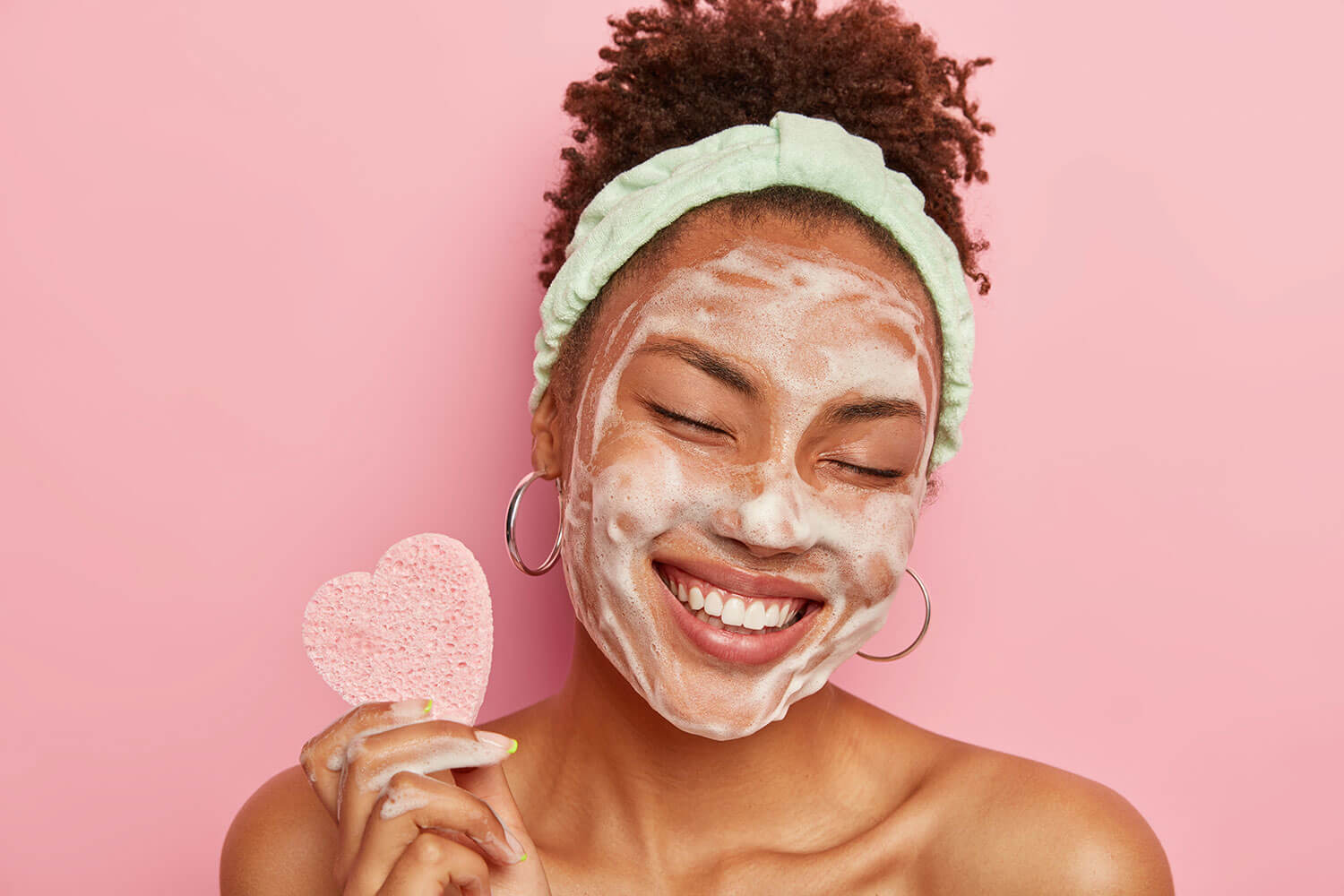 Top 6 Face Washes for Normal Skin. Beautiful optimistic Afro American woman cleanses face with foam, refreshes skin, has well cared complexion, holds heart shaped sponge for beauty procedures, stands bare shoulders with closed eyes.