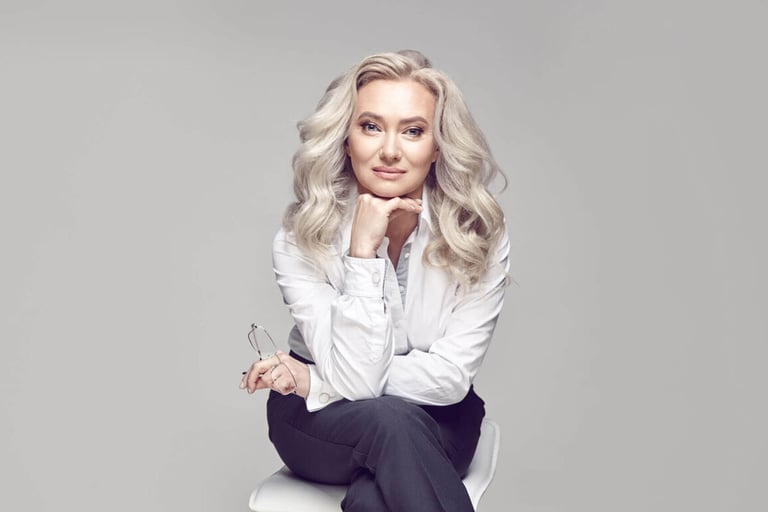 How to Care for Gray Hair. A pretty older woman with beautiful and healthy blonde hair and clear skin, looks beautiful, dressed in a strict and stylish business image, looks at the camera subtly, sits on a chair on a grey isolated background.