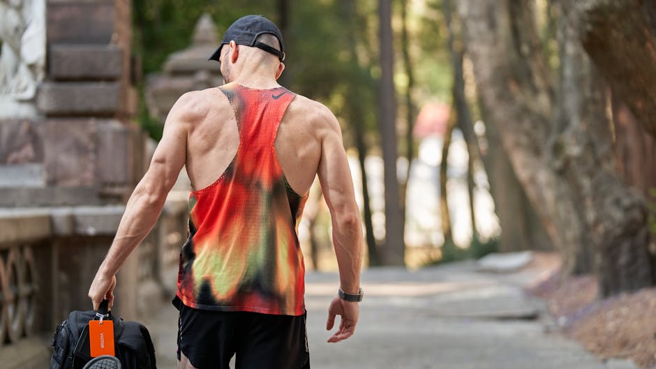 A muscular male runner in a bright tank top carries a gym bag while strolling outdoors.