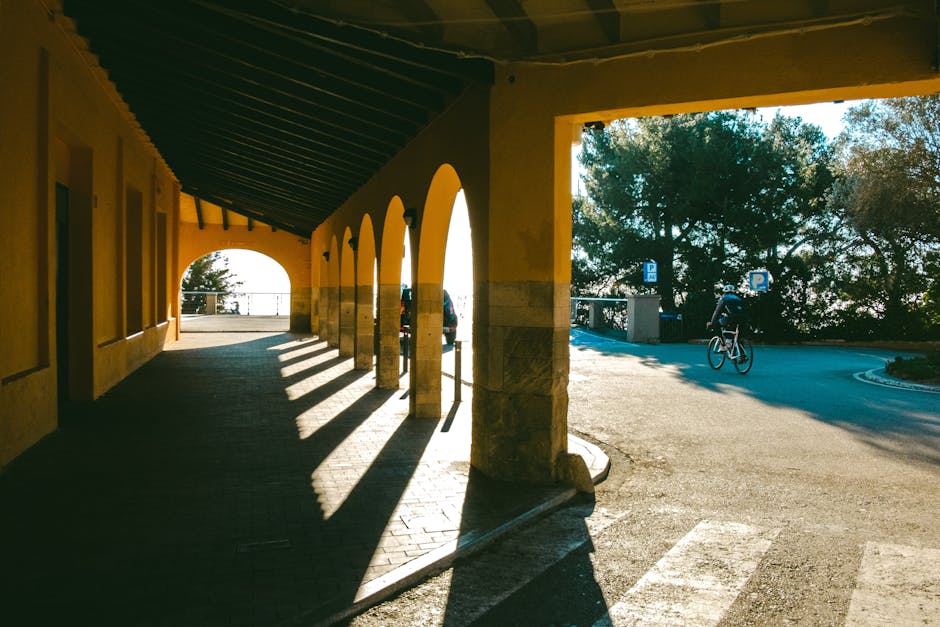 A cyclist rides past a sunlit arched walkway, casting long shadows in a serene urban setting.