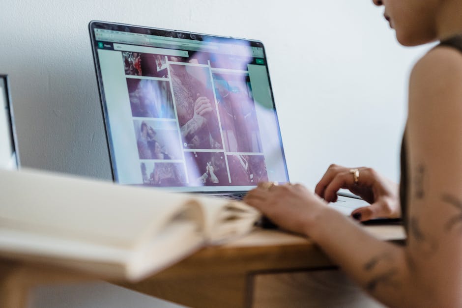 Crop unrecognizable female freelancer with tattoos sitting at table and browsing photos on laptop in room in daytime