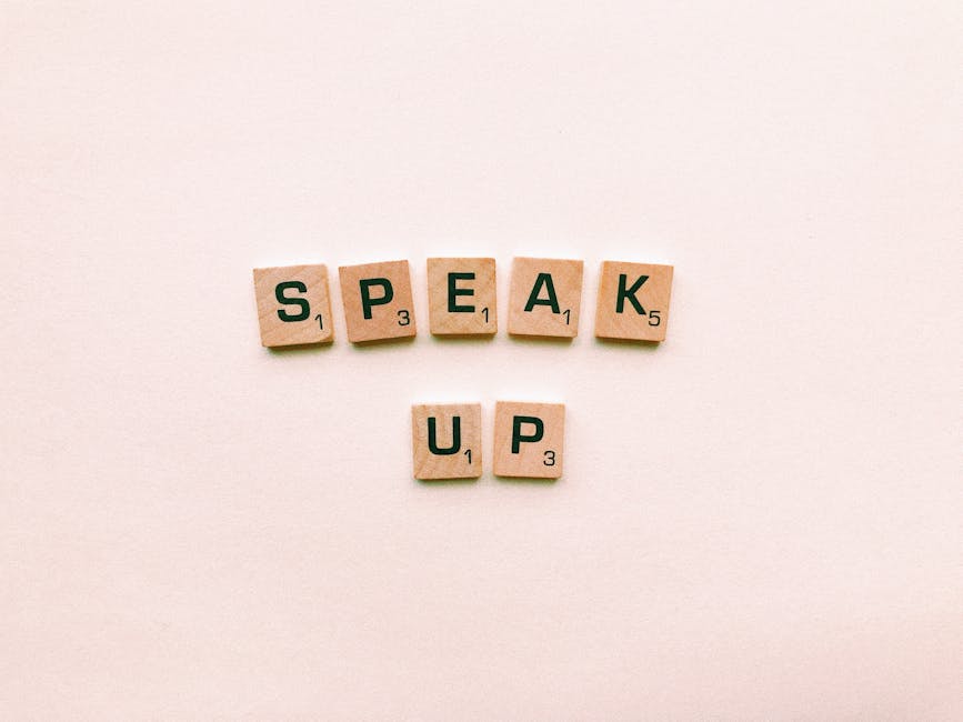 Wooden Scrabble tiles spelling 'Speak Up' on a pink background.