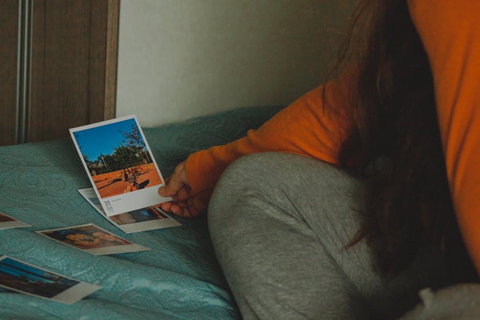High angle of crop anonymous female with long dark hair in comfy clothes sitting on bed and watching instant photos