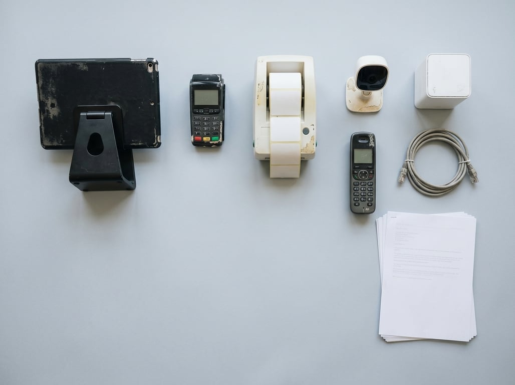 Overhead flat-lay of a tablet on a weighted counter stand, card terminal, label printer with blank labels, indoor camera, mesh wifi node, network cable, cordless handset and printed sheets