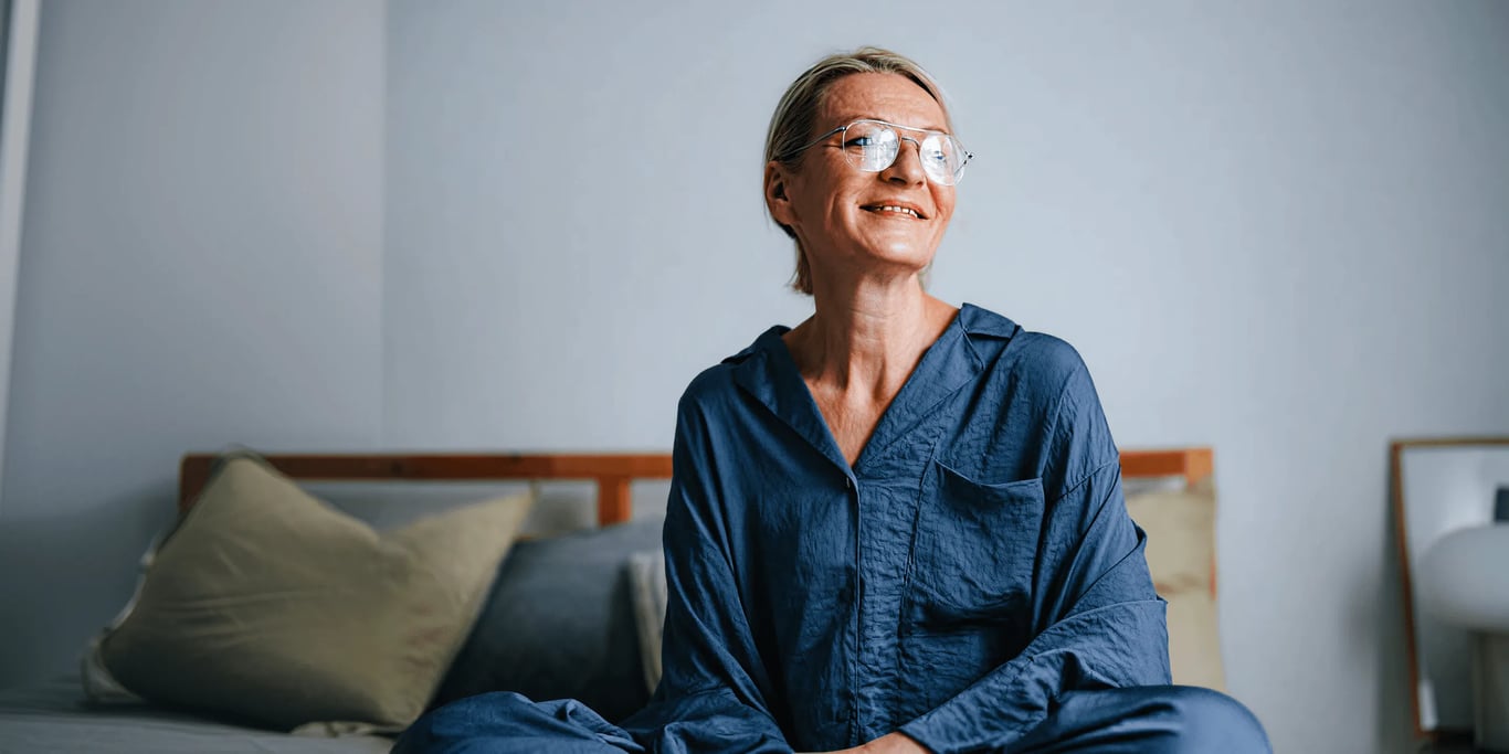 full-width photo of a smiling older woman with glasses in a blue shirt, seated in a bedroom