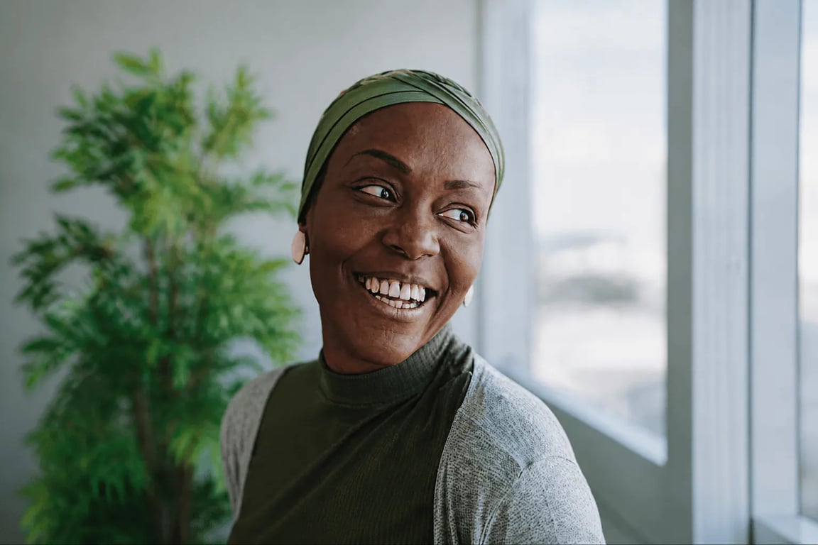 Full-bleed photo of a smiling woman in an olive-green headwrap looking to the side, indoors beside a window with a leafy plant behind her