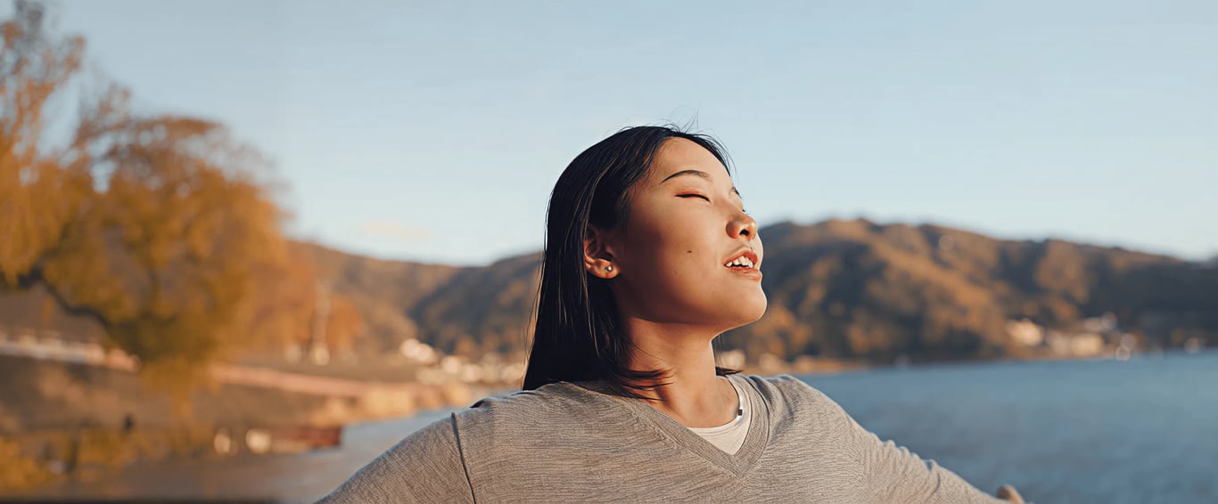 full-width photo of a woman with eyes closed smiling in golden sunlight beside a lake with hills in the background