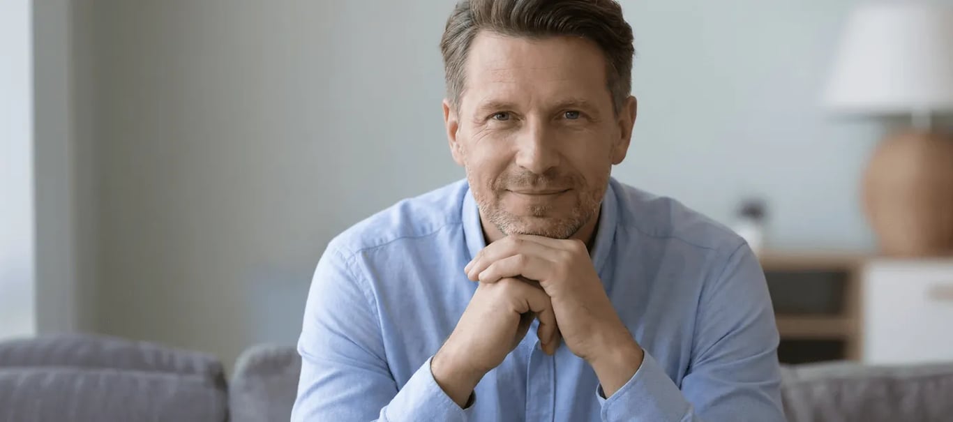 full-bleed photo of a middle-aged man in a light blue shirt smiling with chin resting on clasped hands