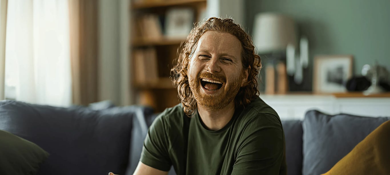 laughing bearded man with curly auburn hair on a couch in a living room