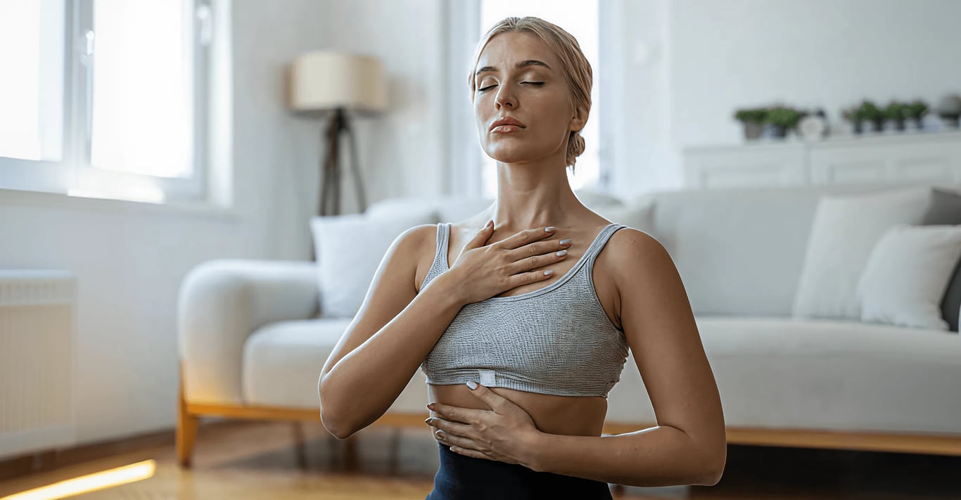 Full-width photo of a woman in a grey sports bra with eyes closed, one hand on her chest and one on her stomach, in a bright living room
